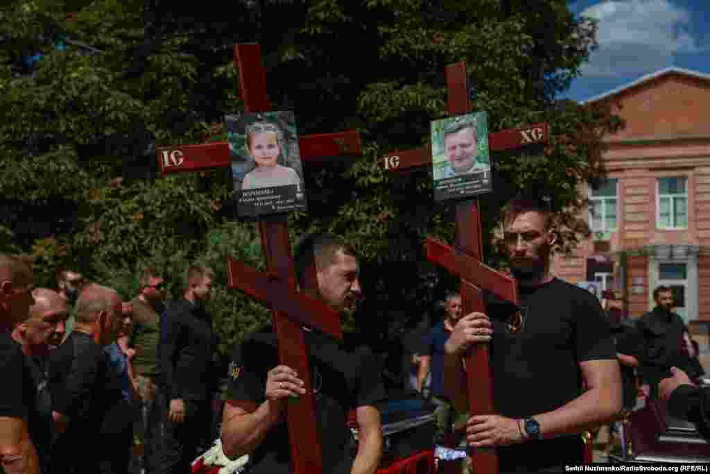 Чоловіки тримають хрести з фотографіями загиблого батька родини Артема Воронова та доньки Емілії. Радушне, Дніпропетровська область, 3 серпня 2026 року.3 серпня у Радушному на Дніпропетровщині прощалися з 5 членами сім'ї Воронових, яких вдалося упізнати: батьками родини – подружжям Артемом та Оленою, а також їхніми трьома дітьми: Марком, Азарієм Ілаєм та Емілією