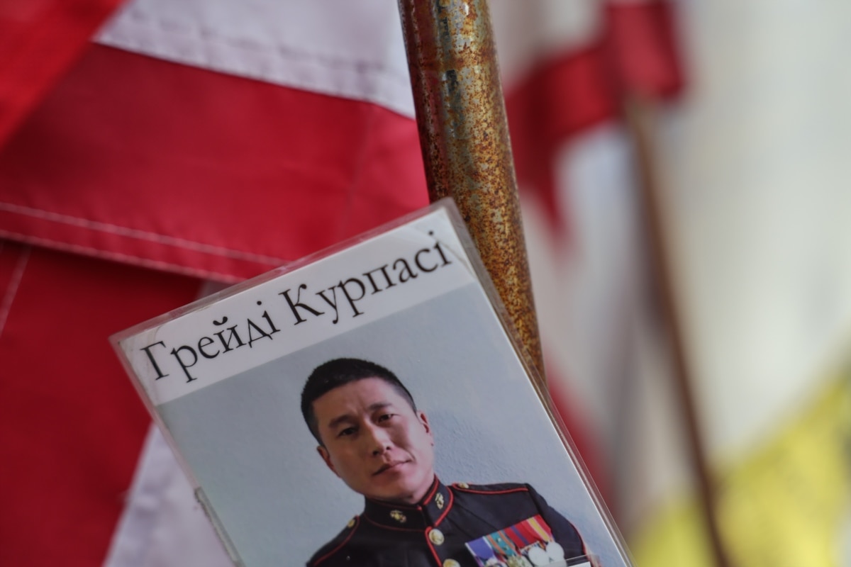 Одним із представників першої хвилі був Грейді Курпасі, колишній капітан морської піхоти США та ветеран війни в Іраку. У 2022 році він приєднався до українських сил і служив у розвідувальному підрозділі Інтернаціонального легіону Team Raven. За повідомленнями з<a href="https://www.rollingstone.com/culture/culture-features/grady-krupasi-marine-ukraine-disappeared-1234765816/" class="wsw__a"> відкритих джерел</a>, Курпасі загинув під час бойових дій на Херсонщині, намагаючись врятувати своїх підлеглих від артилерійського обстрілу. Згодом його перепоховали у США на Арлінгтонському національному цвинтарі.<br><br>На фото: портрет Грейді Курпасі на народному меморіалі пам’яті на майдані Незалежності в Києві, червень 2026 року