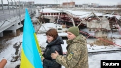 În fundal, Podul Romanivka (Romanivski), simbol al rezistenței ucrainene, principala cale de evacuare a civililor din zona frontului, distrus la începutul războiului. Numit și „Drumul Vieții", a jucat un rol crucial în evacuarea a 40.000 de civili din Bucea, Irpin și Hostomel.