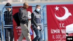 A migrant is escorted by a Turkish police officer walking past a banner reading: "Migration directorate of the Izmir Governor" as they arrive by ferry from the Greek island of Lesbos at the Dikili harbor in Izmir on April 4.