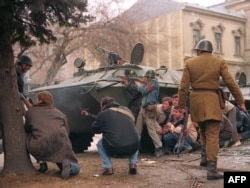Un grup de luptători civili anticomuniști împreună cu un soldat român care-i sprijină pe luptătorii împotriva regimului Ceaușescu se protejează de focul lunetiștilor din spatele unui transport de trupe blindat pe 24 decembrie 1989.