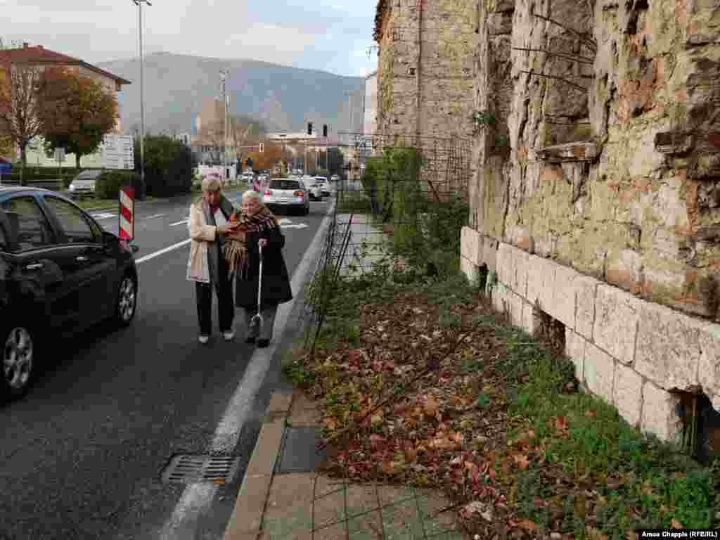 Two elderly women walk into oncoming traffic around a building damaged by war during the early 1990s. 