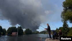 Pescari pe malul unui râu, în timp ce fumul se ridică de la depozitele Wildberries din Koledino, regiunea Moscovei, după un atac cu drone ucrainene, 16 august 2026