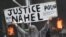 Un participant la proteste ține o pancartă pe care scrie "Justiție pentru Nahel", în timp ce mașinile ard pe stradă la sfârșitul unui marș de comemorare pentru Nahel.