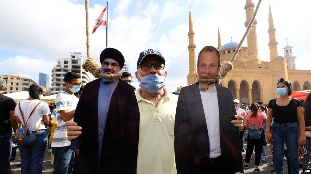A man poses for a picture with a cardboard cut-out of Hassan Nasrallah (L), the head of Lebanon's Hezbollah, and former Foreign Minister Gibran Bassil hung by Lebanese protesters in downtown Beirut on August 8, 2020.