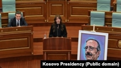 Një fotografi e kongresistit të ndjerë amerikan, Eliot Engel, qëndron para foltorës së Kuvendit të Kosovës, teksa në prapavijë shihen kryeministri Albin Kurti, dhe u.d. e presidentit Albulena Haxhiu, gjatë një seance në nderim të tij, 24 prill.