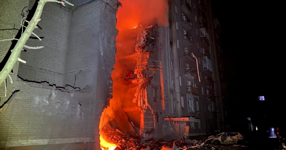 A multi-story apartment building with a large section destroyed and engulfed in fire at night.