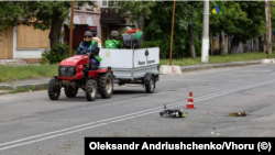 Комунальники у шоломах у Херсоні проїжджають повз збитий російський безпілотник. Червень 2026 року