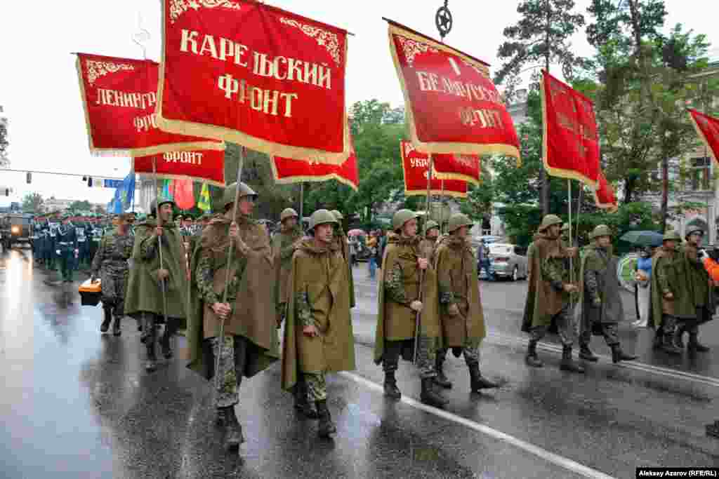 За БТР молодые парни в плащ-палатках и касках времен войны несут штандарты фронтов. Алматы, 9 мая 2016 года.