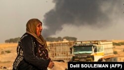 Një grua ecë, teksa në prapavijë shihet tym, pas bombardimeve të Turqisë në qytetin sirian, Rasa l-Ain, që gjendet në kufi me Turqinë.
