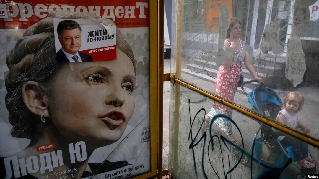 A woman pushes a child on a stroller past an advertisement for a magazine with presidential candidate Yulia Tymoshenko on the cover and an election sticker for current incumbent Petro Poroshenko.
