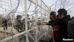 FILE: Refugees and migrants stand next to a border fence at the Greek-Macedonian border.