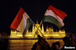 კაცი უნგრეთის დროშებს აფრიალებს, მდინარე დუნაის გადაღმა, საპარლამენტო არჩევნების დღეს. ბუდაპეშტი. 2026 წლის 12 აპრილი