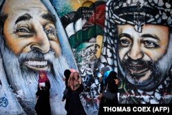 Femei palestiniene trec pe lângă o pictură murală care îi înfățișează pe răposatul lider spiritual al Hamas, șeicul Ahmed Yassin, și pe fostul lider palestinian Yasser Arafat, la 4 mai 2014, în orașul Gaza.
