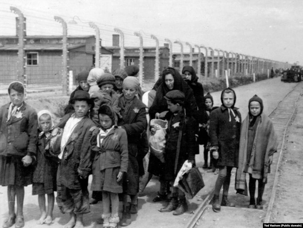 Das rechte Foto stammt aus dem Jahr 1944 und zeigt eine Frau mit ihren Kindern auf dem Weg zur Gaskammer. Dahinter liegen die Holzbaracken des Lagers Birkenau, einem Nebengebäude von Auschwitz. Nach dem Krieg wurden diese Gebäude abgerissen, doch die Ziegelmauern und Schornsteine ​​stehen noch.