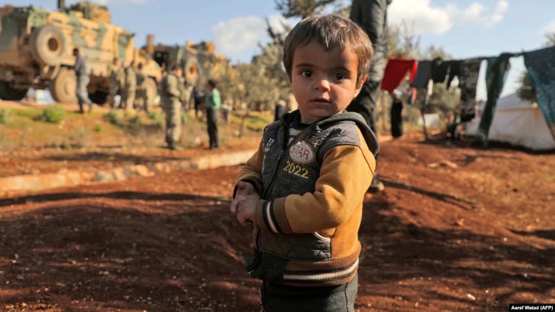 A displaced Syrian child looks to the camera as Turkish soldiers gather on a highway near the war-torn province of Idlib earlier this week. 