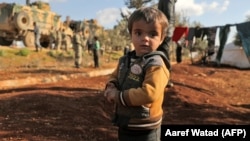 A displaced Syrian child looks to the camera as Turkish soldiers gather on a highway near the war-torn province of Idlib earlier this week. 