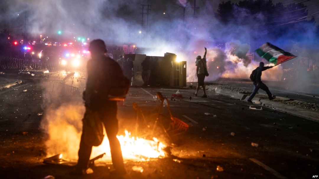 Un protestatar flutură steagul palestinian (în dreapta imaginii) în timp ce forțele de ordine se ciocnesc cu manifestanții în timpul unui protest inn cartierul Compton din Los Angeles, California, pe 7 iunie 2025.