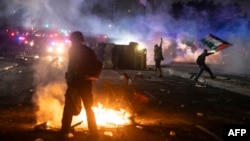 Un protestatar flutură steagul palestinian (în dreapta imaginii) în timp ce forțele de ordine se ciocnesc cu manifestanții în timpul unui protest inn cartierul Compton din Los Angeles, California, pe 7 iunie 2025.