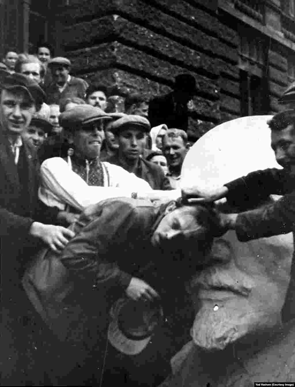 Толпа издевается над евреем возле бюста Ленина на западе Украины. После оккупации нацистские войска открыли тюрьмы советских спецслужб. Преступления сталинизма стали известны и их использовали нацистские пропагандисты, чтобы разжечь антисемитизм, делая упор на еврейском происхождении Сталина.