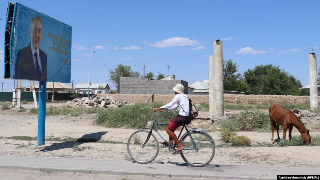 Шорнақ ауылына берісте тұрған бұрынғы президент Нұрсұлтан Назарбаевтың бейнесі бар жарнаманың қасынан велосипед мініп өтіп бара жатқан адам. Түркістан облысы, Сауран ауданы, 30 маусым 2021 жыл.
