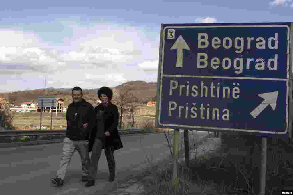 A couple walks past a traffic sign pointing toward the capitals of Serbia and Kosovo.