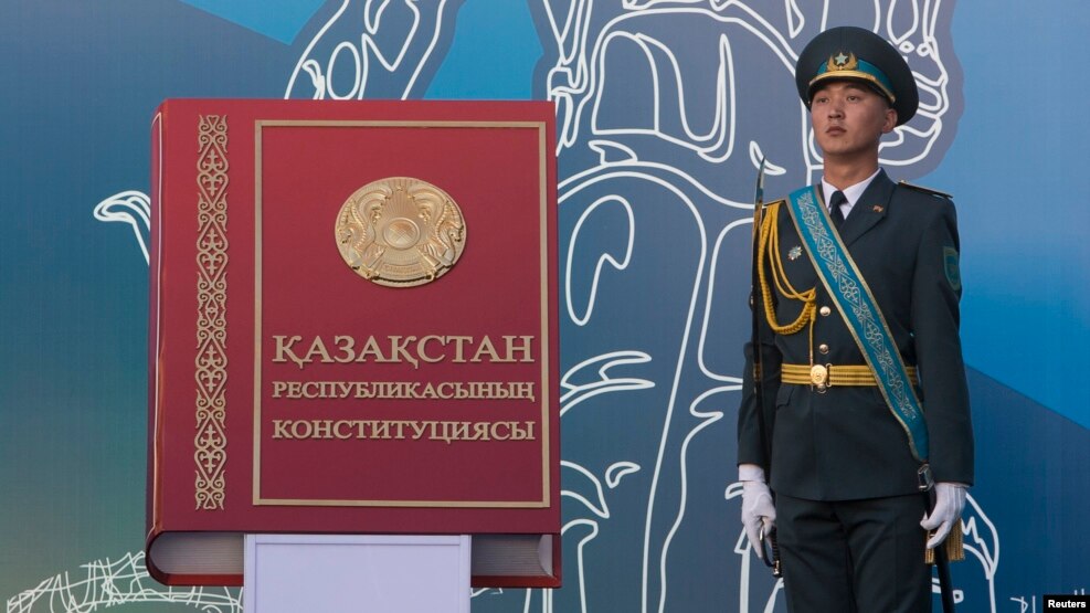 Qazaqstan konstituciyasınıñ kitap türindegi formasın beyneleytin installyaciya men onıñ janında twrğan qarauıl (Körneki suret).