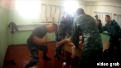 The video shows a prisoner lying on a desk without pants while two people in uniform hold his hands behind his back. At least 10 other uniformed men repeatedly strike his legs and heels with rubber truncheons and fists.