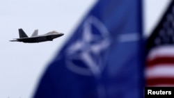Майорять прапори НАТО та США, коли винищувач F-22 Raptor ВПС Сполучених Штатів пролітає над військовою авіабазою в Шяуляї, Литва, 2016 рік