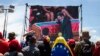 هواداران چاوز، بر پرده‌ای در خیابان، ادای احترام محمود احمدی‌نژاد به رئیس‌جمهور فقیدشان را تماشا می‌کنند.