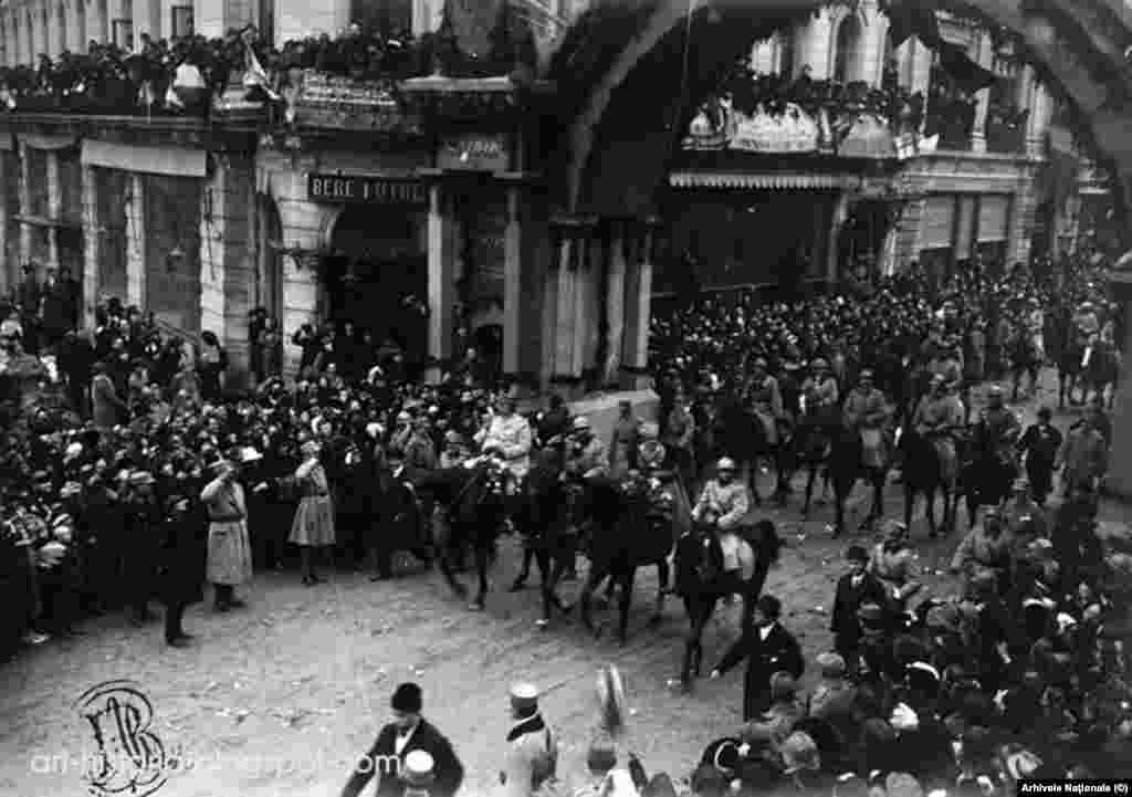 Regele Ferdinand, Regina Maria, flancați de principele moștenitor Carol și generalul Berthelot, trecând pe sub un Arc de Triumf, improvizat pe Calea Victoriei.