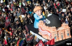 Tilly nu este nicidecum „fixat” doar pe Putin. În imagine, un car alegoric de-al său din 2015, cu cancelarul de atunci al Germaniei, Angela Merkel. Ea nu l-a dat în judecată.