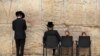 Izraeli gyerekek egy széken ülnek, miközben ultraortodox férfiak a jeruzsálemi Siratófalnál imádkoznak, a fal a judaizmus egyik legszentebb helye.