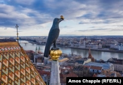 A raven with a ring in its beak looks east over the Danube from its golden perch on the Matthias Church.