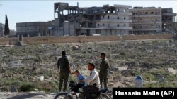 На фото, снятом в марте 2018 года, местный житель с ребёнком и двое курдов из сил безопасности на окраине сирийского города Манбиджа.