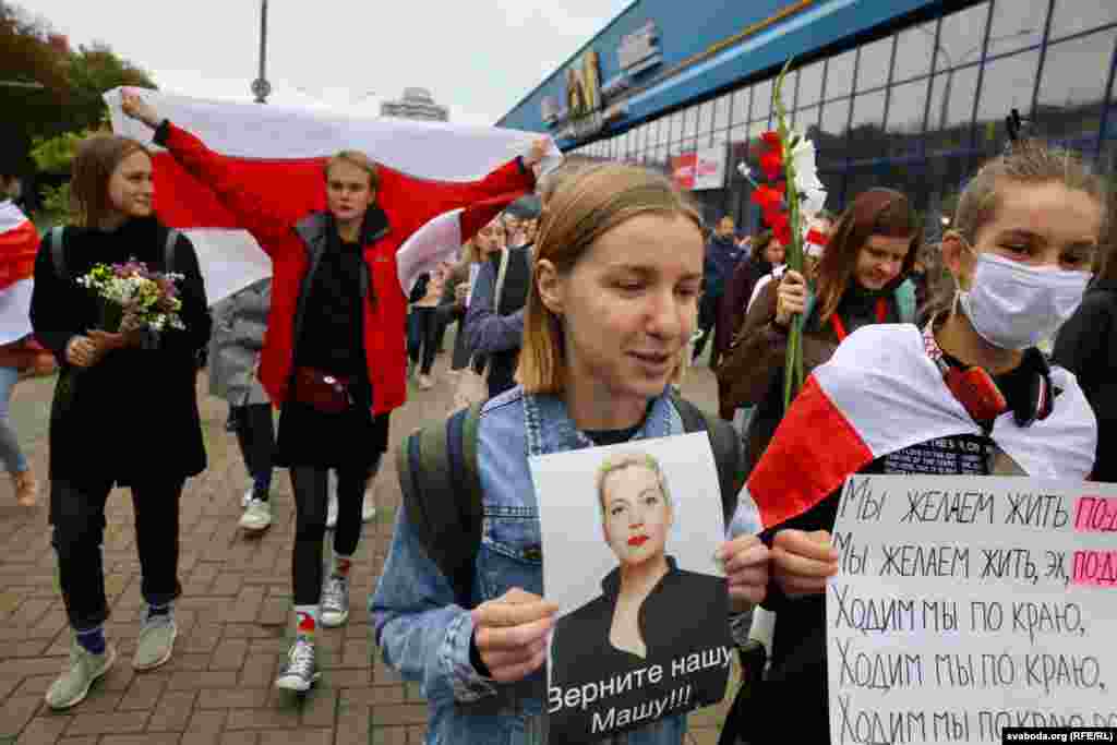Un susținător cu un portret al Mariei Kolesnikova pe care scrie: &bdquo;Dați-ne-o înapoi pe Mașa!!!&quot; în timp ce coloane de protestatari&nbsp;se îndreaptă către Bulevardul Independenței din centrul Minskului. Femeia de lângă ea ține o placardă pe care scrie: &bdquo;Vrem să trăim altfel&rdquo;.