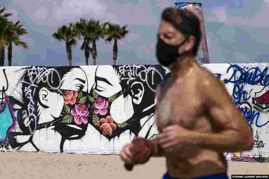 Мъж с маска, тича на плажа край графит, изобразяваш двойка, която се целува през предпазни маски. Графитът е във Венис, Калифорния.