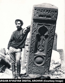 A man leans on a khachkar topped with a winged creature in the Julfa cemetery in 1915.