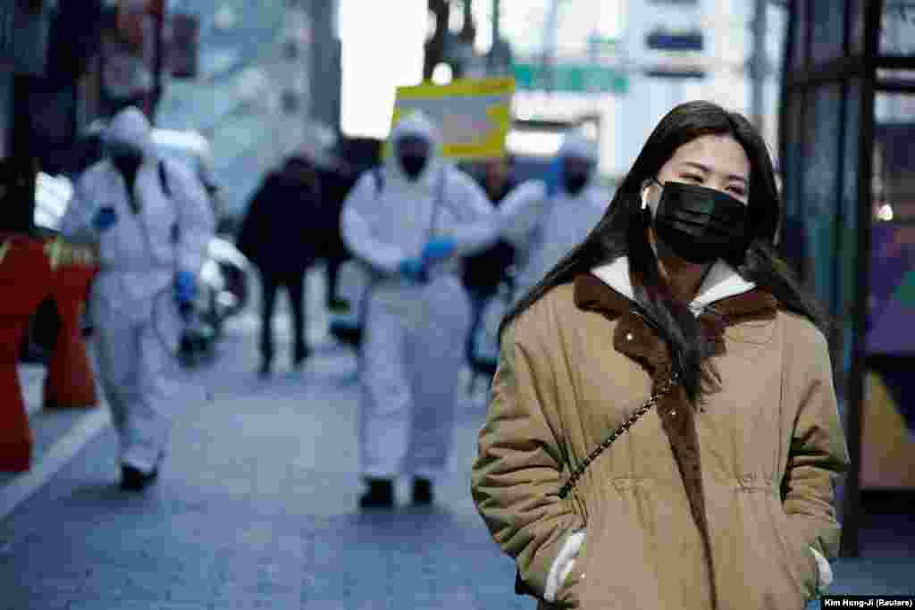 Женщина в маске проходит мимо южнокорейских солдат в защитной одежде, проводящих дезинфекцию улиц Сеула. В Южной Корее коронавирусом заразилось свыше семи тысяч человек, более 50 человек умерли. 5 марта 2020 года.&nbsp;