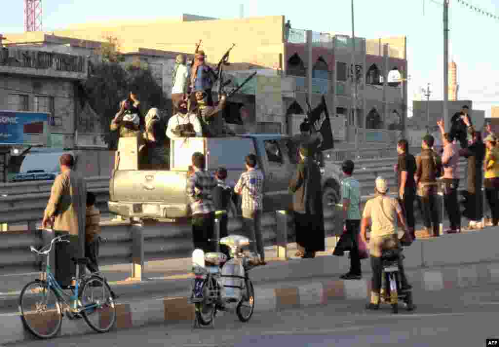 In Fallujah, life goes on. Local&nbsp;Iraqis stand watching an armored vehicle carrying militants holding an Islamic jihadist flag.