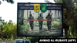 IRAQ -- A picture taken on June 20, 2018 shows a propaganda billboard for the pro-Iran Hezbollah Brigades militia hanging over Palestine Street in the centre of the Iraqi capital Baghdad, depicting three of their masked fighters walking along a road betwe