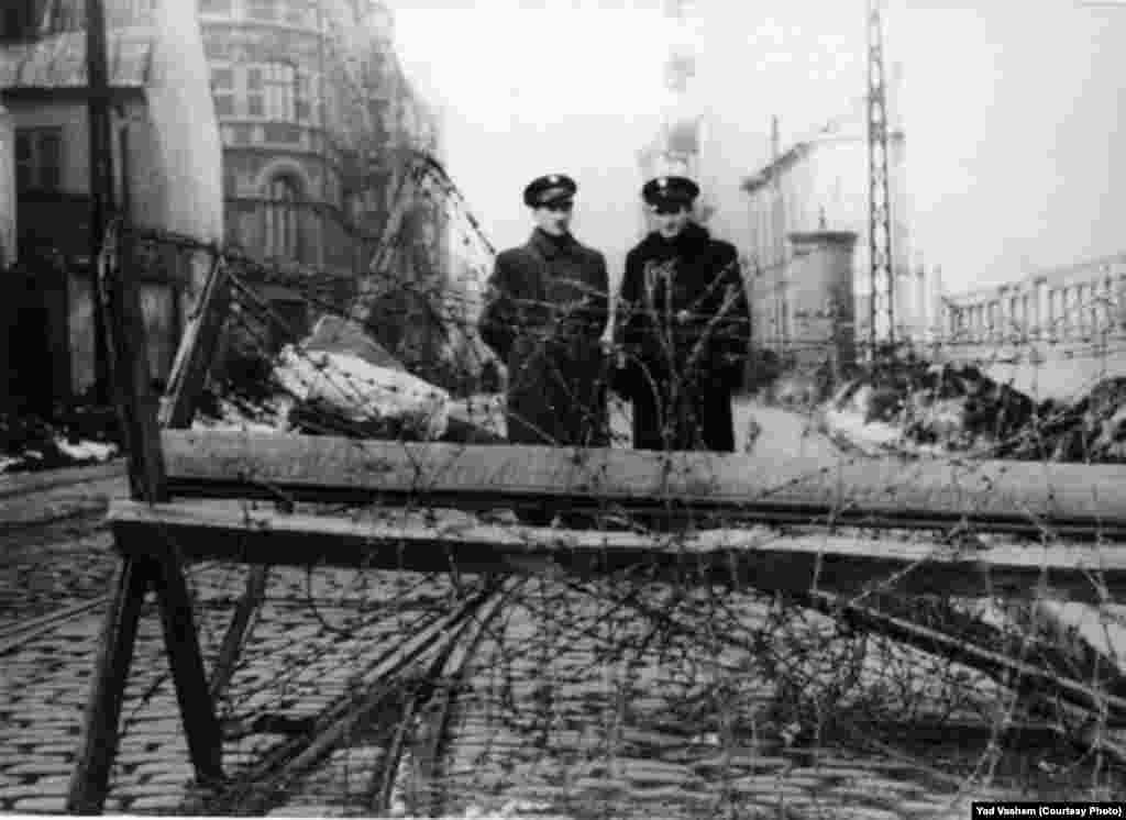 Two Jewish policemen stand near barbed-wire fences.