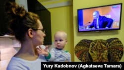 Женщина с ребенком на руках смотрит телевизор, по которому транслируют пресс-конференцию президента России Владимира Путина.