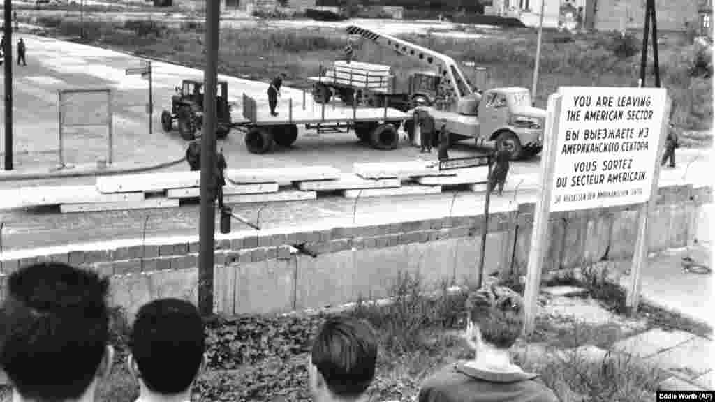 Nyugat-németek (háttal a kamerának) nézik, ahogy kelet-német munkások előregyártott betonlemezeket raknak ki a berlini fal megerősítésére 1961. szeptember 12-én.&nbsp;