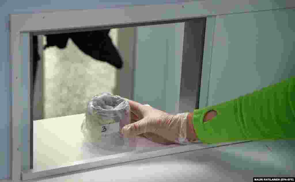 Un eșantion de testare este transmis supraveghetorilor aflați într-o cameră separată. Câinii care detectează coronavirusul încep o perioadă de test de patru luni pe aeroportul Helsinki-Vantaa de lângă Helsinki, 23 septembrie 2020. Ajuns la un punct de eșantionare în aeroport, un pasager intră într-o rezervă, colectează un eșantion de pe suprafața pielii și îl pune într-un container conform instrucțiunilor. Câinele și îngrijitorul său așteaptă în spatele unui perete unde câinele adulmecă eșantionul.&nbsp;