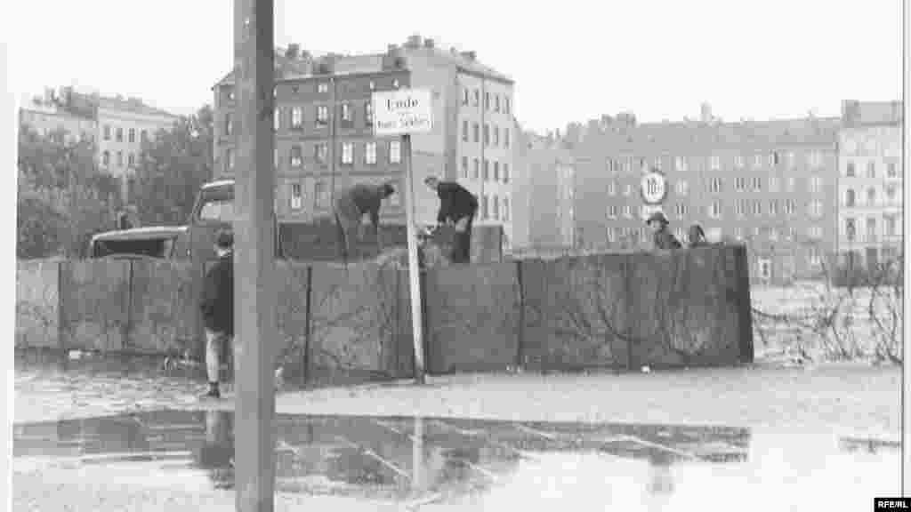 Kelet-német katonák őrködnek az egy éjszaka alatt felhúzott fal egyik átmeneti pontján. Az akkori kelet-német vezető, Walter Ulbricht két hónappal azelőtt, június 15-én még azt mondta &quot;Senki sem akar falat építeni&quot; a városban. egy hónappal később azzal indokolta a fal megépítését, hogy az&nbsp; egy &quot;antifasiszta védmű.&quot; &nbsp;
