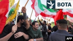 Disa persona, duke mbajtur flamuj të Iranit, Libanit dhe shteteve të tjera, festojnë para Ambasadës iraniane në Bagdad për arritjen e marrëveshjes mes Iranit dhe SHBA-së. 15 qershor 2026. 

