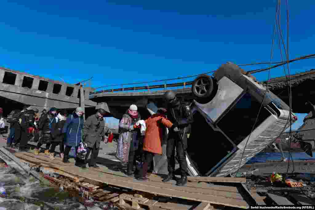 Поліцейський допомагає цивільним, які покидають Ірпінь, переходити річку по дошках поруч із підірваним мостом. Межа між Ірпенем і житловим масивом Романівка, 11 березня 2022 року