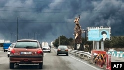 Nori groși de fum pe cerul Teheranului, în urma unor noi lovituri americane și israeliene, pe 8 martie 2026, văzuți de pe o stradă dominată de o statuie în cinstea victoriilor iraniene în războaie din trecut și de un panou cu imaginea fostului lider suprem ucis, Ali Khamenei