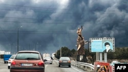 Makina kalojnë pranë një statuje përkujtimoreqë paraqet udhëheqësin suprem të vrarë të Iranit, Ajatollah Ali Khamenei, më 8 mars në Teheran, ndërsa shtëllunga tymi nga sulmet ndaj objekteve të naftës mbulojnë qytetin.